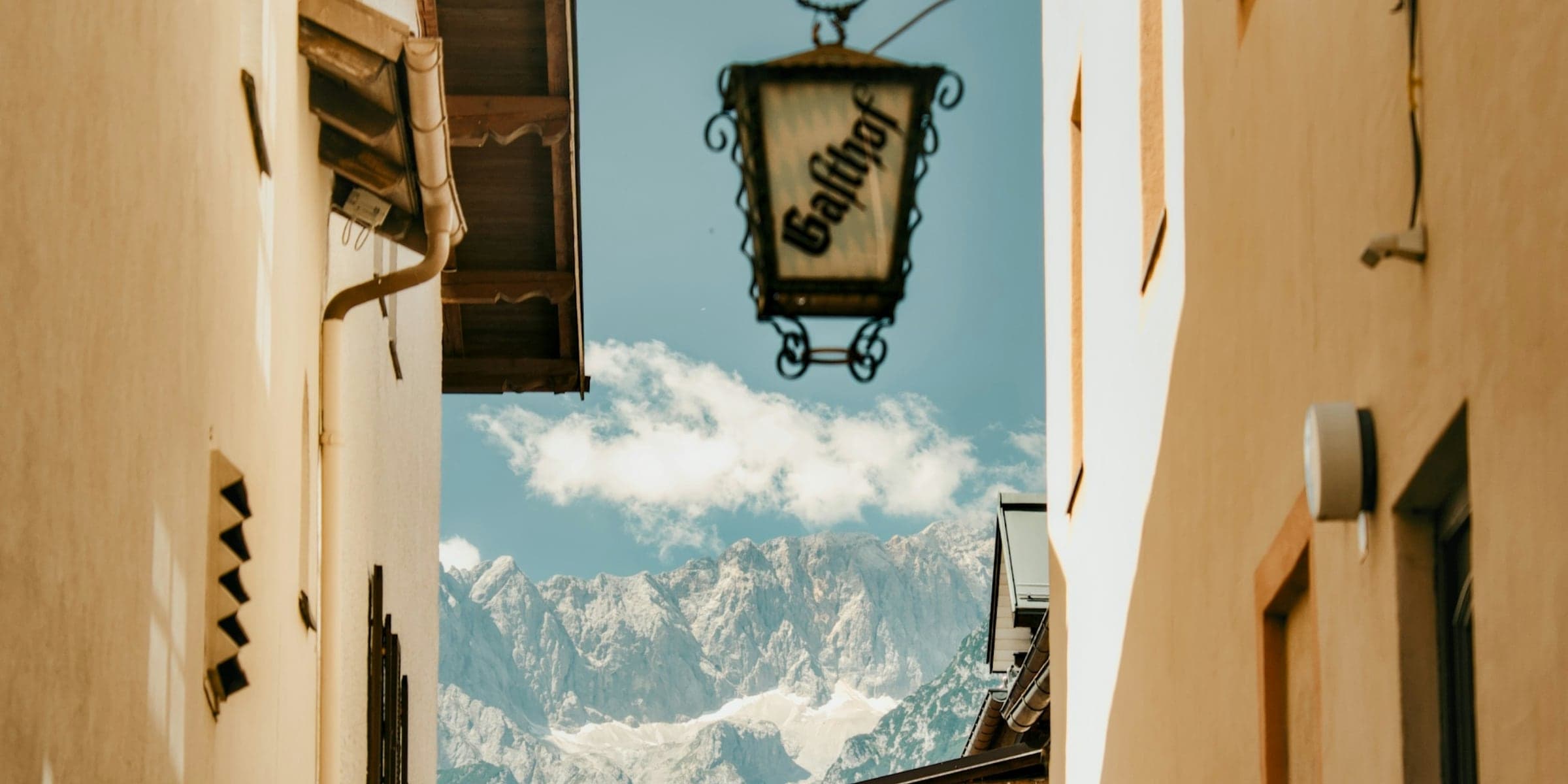 A hand-forged inn lantern in a narrow alpine lane, the mountain standing at the end of it.